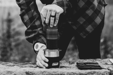 A black and white picture of a person outside, using an AeroPress travel coffee maker. Brewing coffee directly into a camp mug.