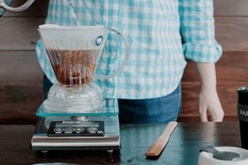 A person in a blue checkered shirt and blue jeans pouring water into a Clever dripper coffee brewer that is sitting on a scale