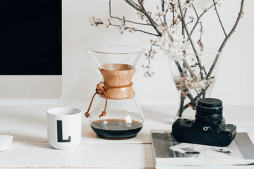 Glass Chemex with a wooden collar sitting on a white desk next to a black camera and a white ceramic mug.